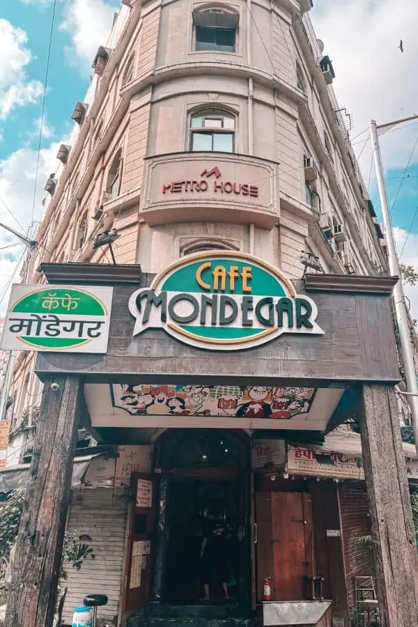 Exterior view of Cafe Mondegar at Metro House on Colaba Causeway, with the cafe's classic Art Deco signage in English and Marathi, and Mario Miranda murals visible above the entrance
