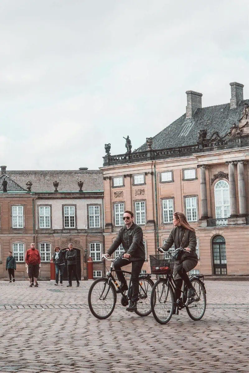 a couple riding bicycles through a cobbled square in copenhagen, denmark, with historic buildings and curious onlookers in the background