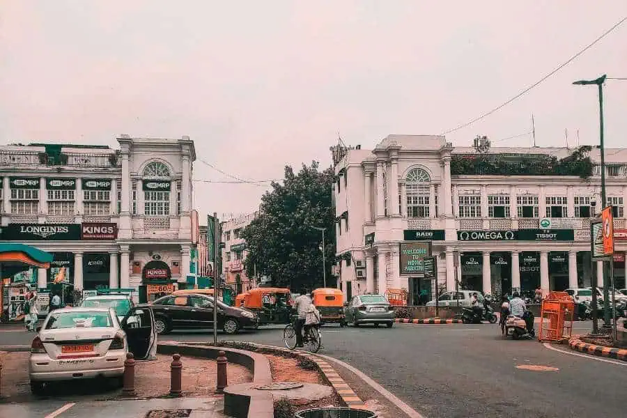 a busy street scene at connaught place, delhi, with colonial-era white buildings housing brand stores like omega and tissot, auto-rickshaws, cars, and cyclists navigating a traffic circle under an overcast sky