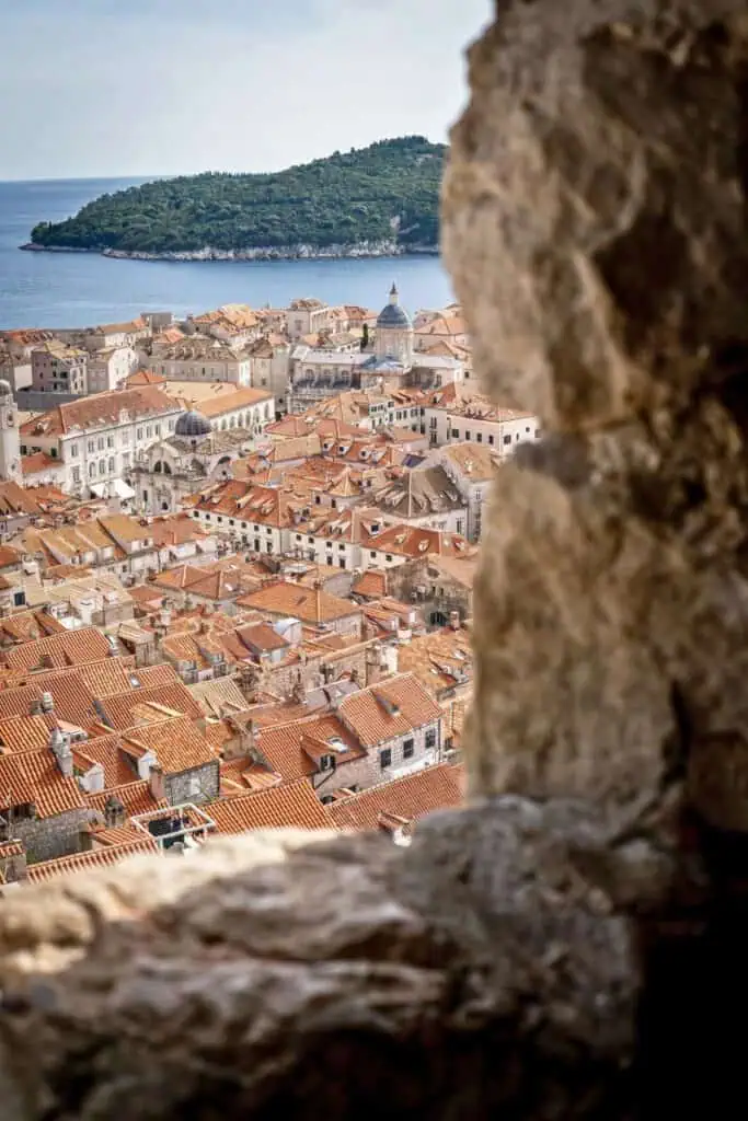 view of dubrovnik's old town with terracotta rooftops and domed churches framed through a stone window, with lokrum island and the adriatic sea in the background