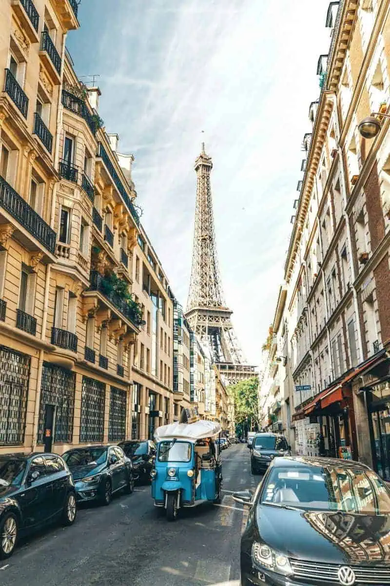 a narrow Parisian street lined with classic Haussmann-style buildings, with the Eiffel Tower rising in the distance and a blue tuk-tuk driving between parked cars