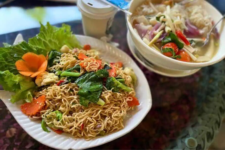 stir-fried noodles with fresh vegetables, garnished with a carved carrot flower, served alongside a bowl of noodle soup at lalitta cafe in chiang rai, thailand