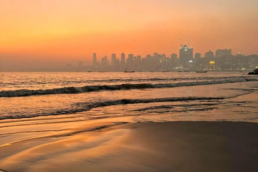 Sunset at Girgaon Chowpatty Beach in Mumbai, with traditional Koli fishing boats anchored in the calm sea and the city skyline silhouetted against an amber sky
