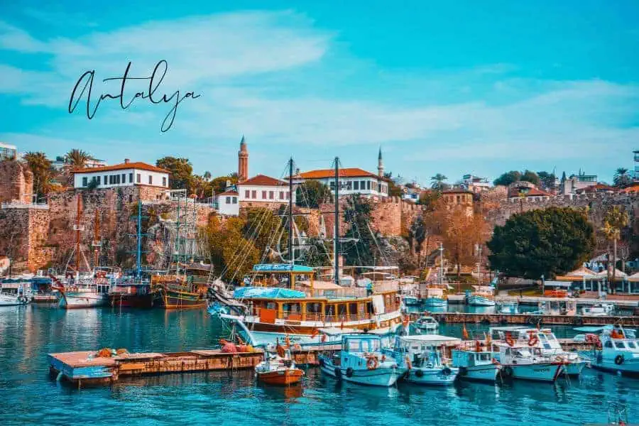 colorful boats docked in the old harbor of antalya, with historic stone buildings and a mosque minaret in the background under a bright blue sky