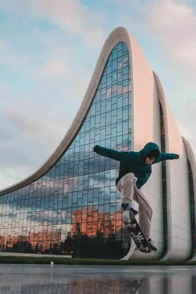 a skateboarder mid-jump in front of the futuristic heyday aliyev center in baku, azerbaijan, with its iconic curved architecture reflecting the cityscape at sunset
