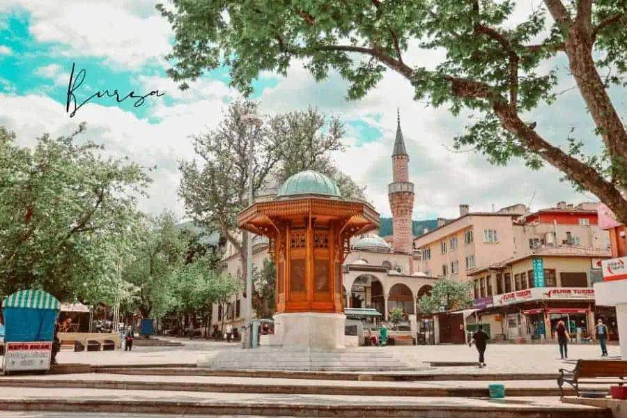 a lively square in bursa with a traditional ottoman-style fountain, surrounded by trees, a mosque minaret, and historic buildings
