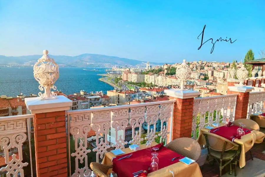  a scenic view of izmir’s coastline from a terrace restaurant, overlooking the blue aegean sea with the city skyline and mountains in the background