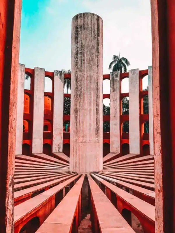 close-up of one of the astronomical instruments at jantar mantar in delhi, showing precise geometric design with symmetrical arches, red-and-white stone structure, and a tall cylindrical pillar at the center against a partly cloudy sky
