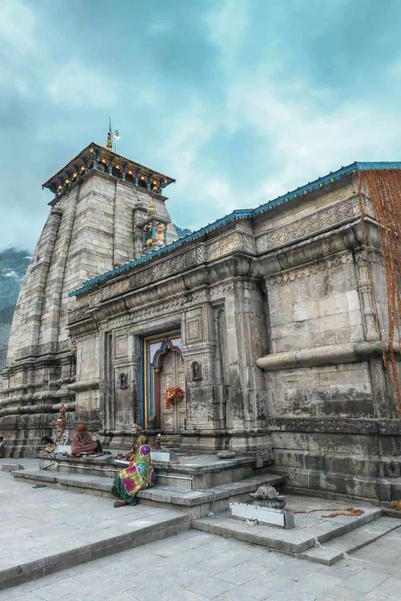 front view of kedarnath temple in uttarakhand, india, with pilgrims sitting near the entrance against a backdrop of mountains and cloudy skies