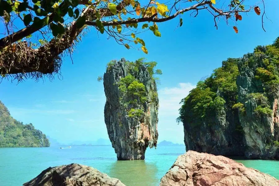 "james bond island (koh tapu) in phang nga bay, phuket, featuring a towering limestone rock formation surrounded by emerald green waters and lush cliffs, framed by tree branches