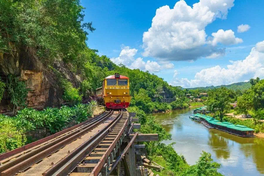 A yellowand red  train crossing a river on the death railway near the river kwai  in kanchanaburi in thailand with green scenery and a blue sky
