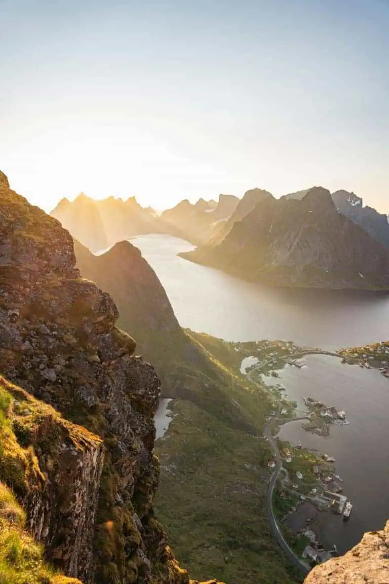 dramatic aerial view of norway’s rugged coastline and towering mountains bathed in golden sunlight, with a winding road leading through a coastal village by the fjord