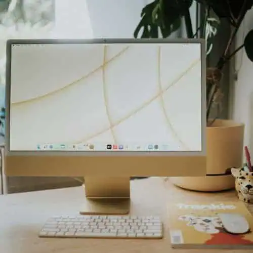 pastel green Apple iMac on a wooden desk with wireless keyboard, next to a plant and a magazine