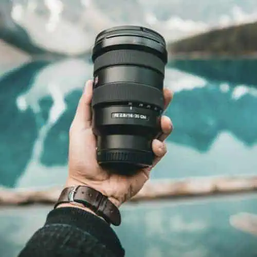 person holding a Sony FE 16-35mm camera lens with a scenic lake and mountains blurred in the background