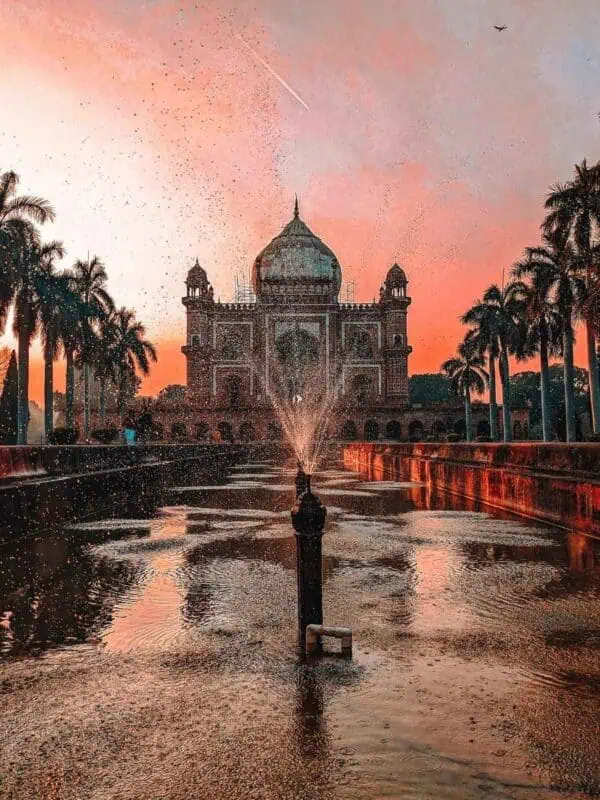 low-angle view of safdarjung tomb in delhi captured at sunset with water droplets frozen mid-air from a central fountain, flanked by palm trees and a warm sky streaked with light