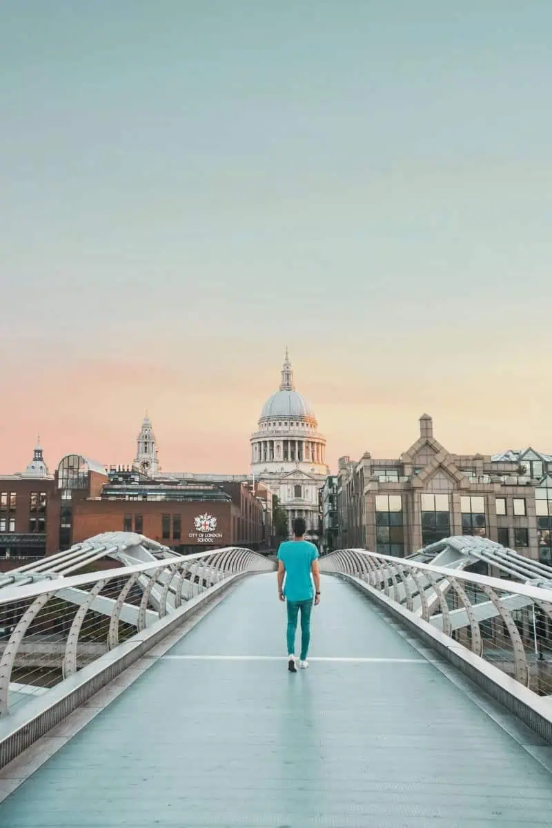 a person walking alone on the Millennium Bridge in London at sunrise, with a clear view of St. Paul's Cathedral framed by modern and historic buildings under a soft pastel sky