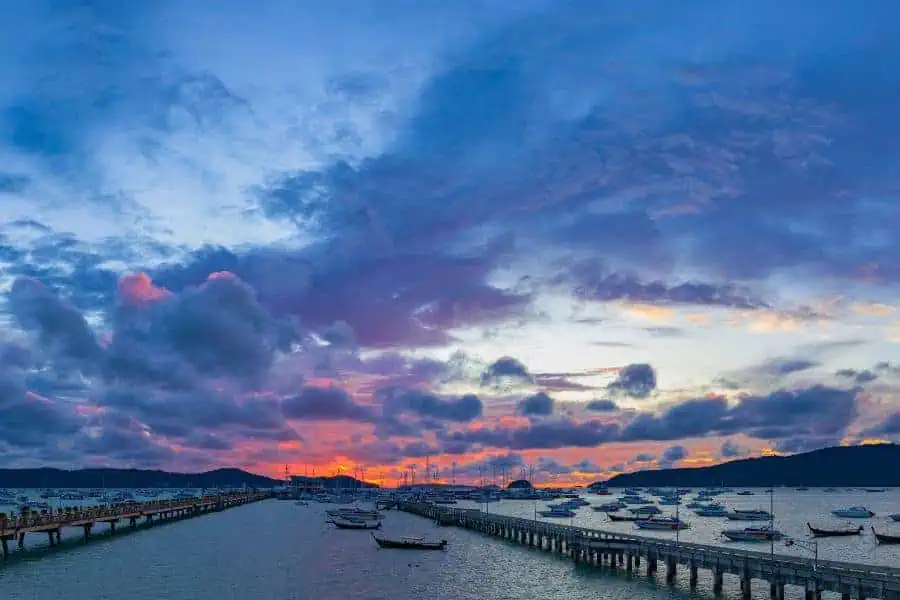 "sunset over chalong pier in phuket with colorful clouds, boats anchored in the bay, and a wooden pier stretching into the calm waters
