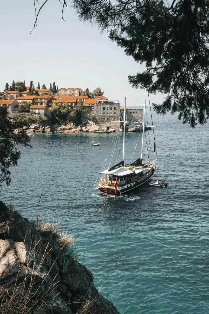 cenic view of a sailboat on turquoise water near the coastline of sveti stefan, montenegro, with red-roofed stone buildings and trees in the background