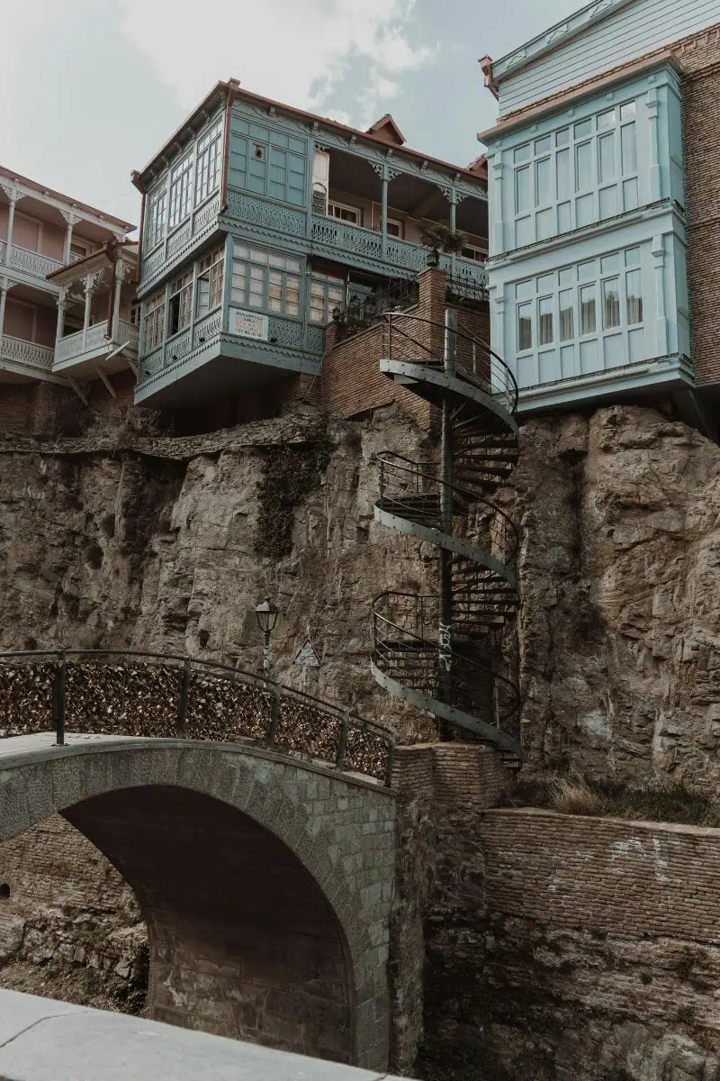 wooden blue balconies of traditional Tbilisi houses perched on a rocky cliff with a spiral staircase and love lock bridge in the foreground