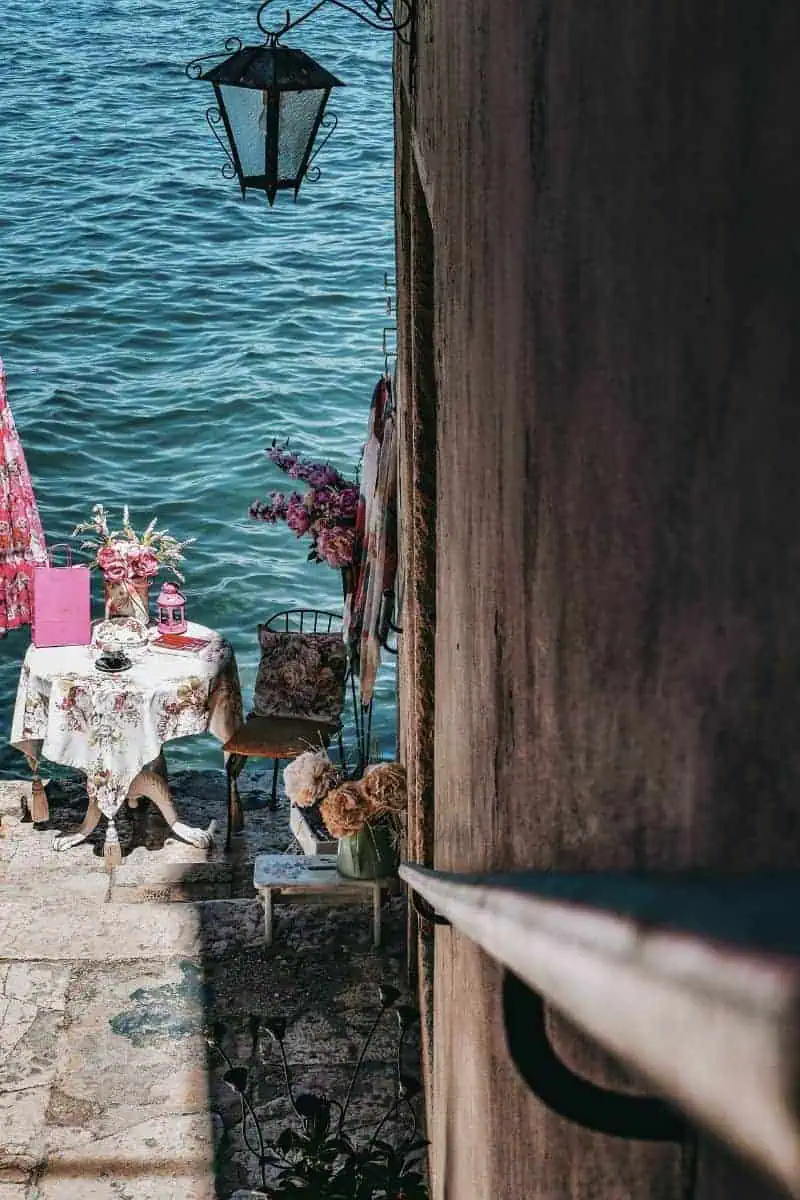 a charming waterfront café corner in Rovinj, Croatia, featuring a floral tablecloth, tea set, and decorative pink details, set against a stone wall overlooking the calm blue Adriatic Sea