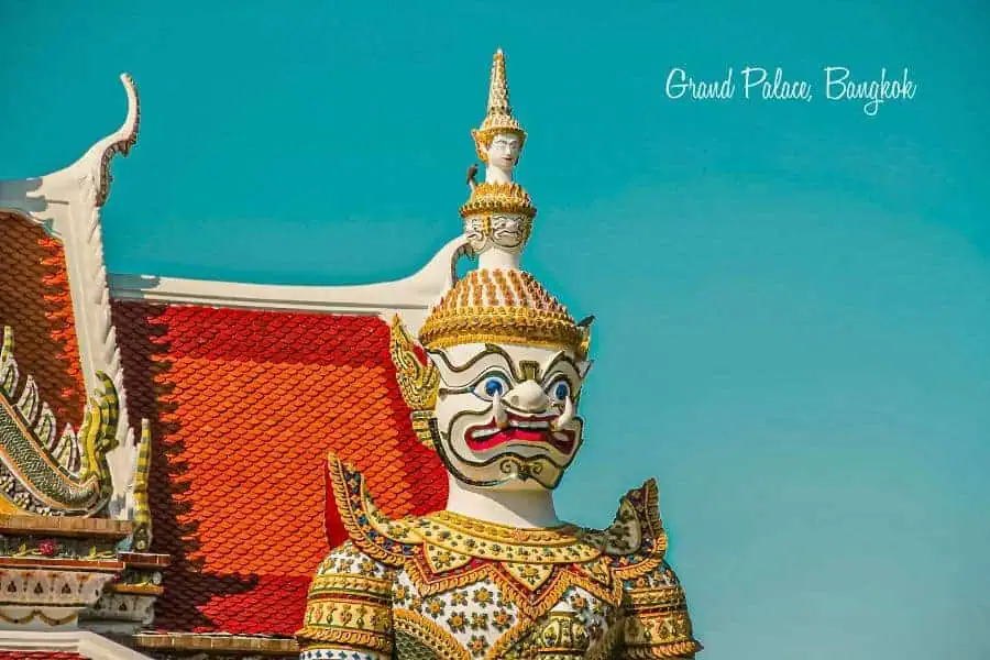 yaksha guardian statue at the grand palace in bangkok, with an ornate orange temple roof and clear blue sky in the background